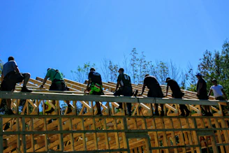 A construction site showing workers carefully assembling the wooden frame of a house under a bright blue sky.