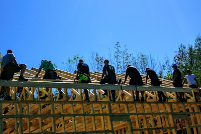A group of construction workers are building a wooden roof structure on a sunny day. The workers, equipped with tools, are actively engaged in the construction under clear blue skies. Surrounding the site is a line of trees, which frames the image.
