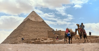 A large stone pyramid towering against a backdrop of partly cloudy blue sky. Several people are walking around the area, with two individuals riding camels in the foreground. The scene conveys a sense of historical grandeur.