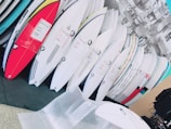 A collection of surfboards neatly organized, with various colors and designs, in what appears to be a store or showroom. The surfboards have protective wrapping and are leaning against a wall. There is bubble wrap on the floor, suggesting the surfboards may be newly delivered or prepared for shipping.