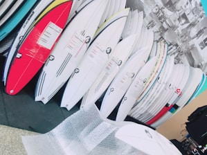A collection of surfboards neatly organized, with various colors and designs, in what appears to be a store or showroom. The surfboards have protective wrapping and are leaning against a wall. There is bubble wrap on the floor, suggesting the surfboards may be newly delivered or prepared for shipping.