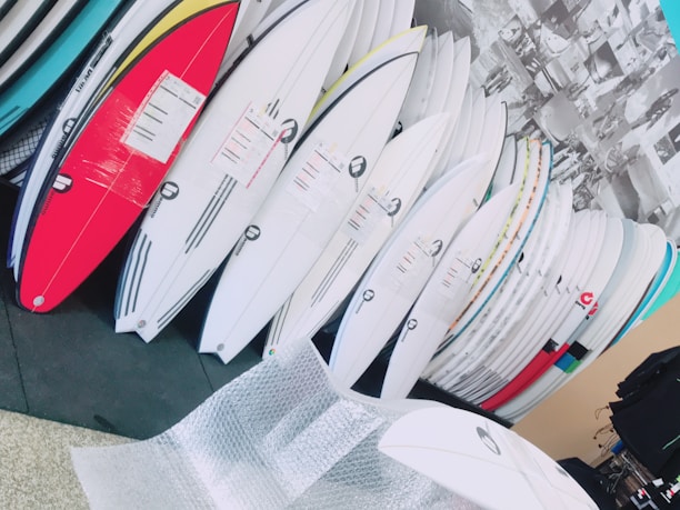 A collection of surfboards neatly organized, with various colors and designs, in what appears to be a store or showroom. The surfboards have protective wrapping and are leaning against a wall. There is bubble wrap on the floor, suggesting the surfboards may be newly delivered or prepared for shipping.