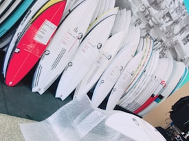 A collection of surfboards neatly organized, with various colors and designs, in what appears to be a store or showroom. The surfboards have protective wrapping and are leaning against a wall. There is bubble wrap on the floor, suggesting the surfboards may be newly delivered or prepared for shipping.
