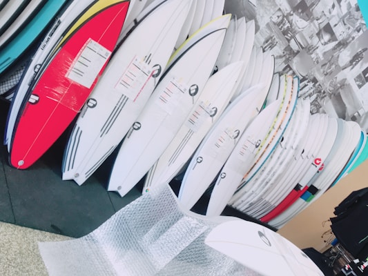 A collection of surfboards neatly organized, with various colors and designs, in what appears to be a store or showroom. The surfboards have protective wrapping and are leaning against a wall. There is bubble wrap on the floor, suggesting the surfboards may be newly delivered or prepared for shipping.