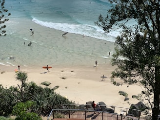 A sandy beach with several people carrying surfboards and walking towards the ocean. Some surfers are already in the water, enjoying small waves. The scene is surrounded by lush greenery and a wooden boardwalk is visible in the foreground, where a couple of people are standing and observing the beach. The sun casts bright light over the landscape, creating a serene and vibrant atmosphere.
