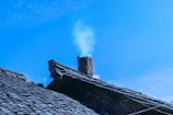 A rooftop with traditional dark tiles and a brick chimney emitting a faint trail of smoke against a clear blue sky.