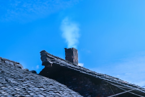 A rooftop with traditional dark tiles and a brick chimney emitting a faint trail of smoke against a clear blue sky.