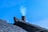 A rooftop with traditional dark tiles and a brick chimney emitting a faint trail of smoke against a clear blue sky.