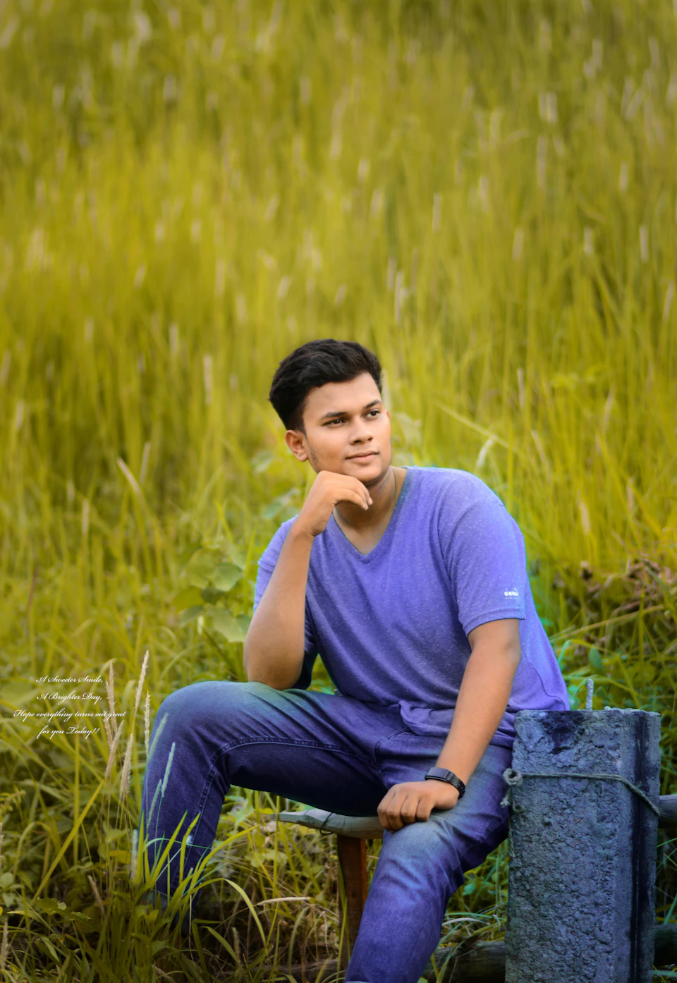 The image shows a young man sitting on a wooden bench, dressed in a blue shirt and jeans. He is looking off to the side with his hand resting on his chin. The setting appears to be an outdoor location, possibly a park or garden, with tall grass surrounding him.