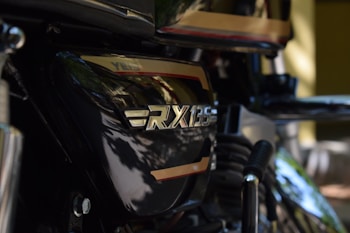 Close-up view of a motorcycle with the RX 135 emblem prominently displayed. The image captures the black and gold accents, with parts of the engine and exhaust pipe visible.