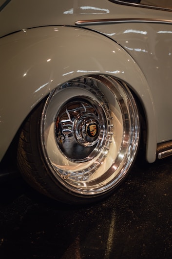 A close-up view of a shiny, polished car wheel with a prominent logo at the center. The surrounding car body is smooth, reflecting light in a garage or showroom setting.
