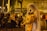 A person dressed in a veil and holding a plush toy is performing on the street with a microphone. A group of people sitting on a curb nearby are watching, and some are smiling. The scene is set at night, illuminated by streetlights with a backdrop of urban buildings.