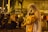 A person dressed in a veil and holding a plush toy is performing on the street with a microphone. A group of people sitting on a curb nearby are watching, and some are smiling. The scene is set at night, illuminated by streetlights with a backdrop of urban buildings.