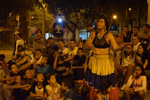 A woman in colorful attire performs with a microphone in hand, surrounded by a large crowd of children and adults seated outdoors in a park setting. There is a mix of eagerness and entertainment evident in the group's expressions, with some onlookers appearing to be taking photos.