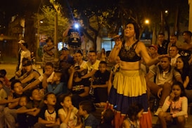 A woman in colorful attire performs with a microphone in hand, surrounded by a large crowd of children and adults seated outdoors in a park setting. There is a mix of eagerness and entertainment evident in the group's expressions, with some onlookers appearing to be taking photos.