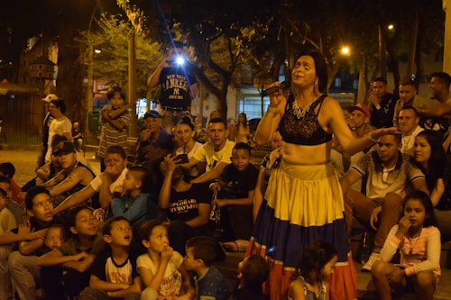 A woman in colorful attire performs with a microphone in hand, surrounded by a large crowd of children and adults seated outdoors in a park setting. There is a mix of eagerness and entertainment evident in the group's expressions, with some onlookers appearing to be taking photos.