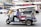 A tuk-tuk is parked on a busy city street. The driver wears a blue shirt and is seated inside the vehicle, which has colorful accents and an ornamental roof. In the background, blurred motion of vehicles and people emphasize the urban setting with multiple shops and signage.