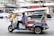 A tuk-tuk is parked on a busy city street. The driver wears a blue shirt and is seated inside the vehicle, which has colorful accents and an ornamental roof. In the background, blurred motion of vehicles and people emphasize the urban setting with multiple shops and signage.