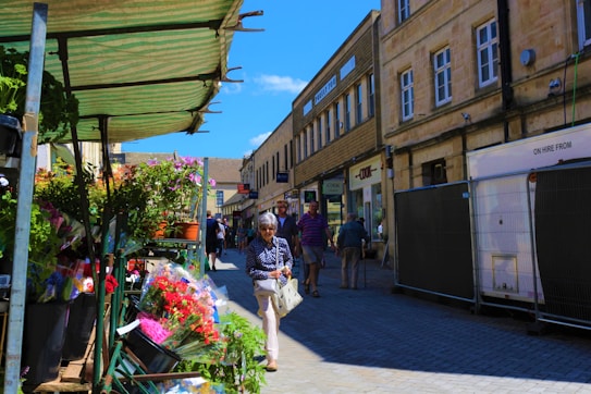 An outdoor market scene with a variety of colorful flowers displayed under a green-striped canopy. People are walking along the street beside stone buildings, which have shops with signs visible. The sky is clear and blue, adding a vibrant and cheerful atmosphere.