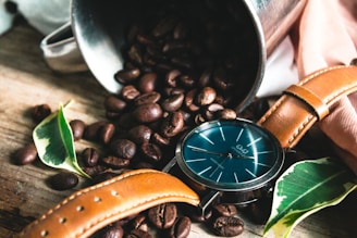 A close-up of a sleek, minimalist wristwatch resting on a wooden table beside a leather-bound notebook and a cup of coffee.