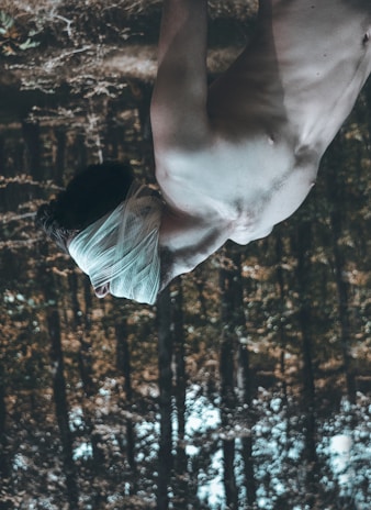 A shirtless person with their head wrapped in gauze, standing in a forest. The image is upside down, creating a surreal effect with trees in the background.