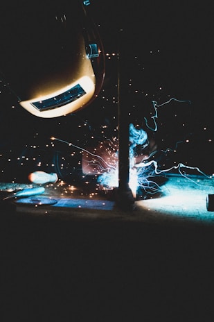 A person is welding a piece of metal, with bright blue and orange sparks flying from the welding point. The person is wearing a protective helmet, and the background is dark, highlighting the glow from the welding process.