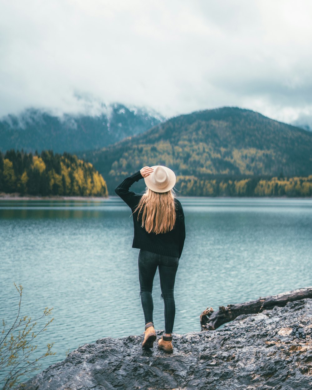 woman in white hat, black jacket, and blue jeans standing near body of water
