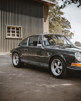 A classic Porsche car is parked beside a wooden building with vertical green panels. The car has a sleek, shiny black finish with prominent detailing on the sides and stylish rims. Sunlight casts soft shadows around the car, enhancing its vintage look. Nearby trees are visible, adding a touch of nature to the scene.