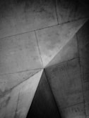 Geometric patterns of intersecting concrete surfaces form sharp angles and lines, creating a modern and abstract architectural detail. The texture of the concrete is visible, adding depth and interest to the composition.