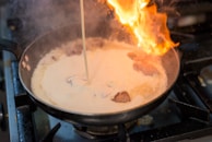 A flaming pan on a stovetop, containing a creamy sauce with visible chunks of food. A stream of liquid is being poured into the pan, causing a flare-up of fire.