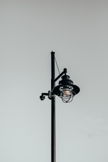 A solitary black street lamp with a modern, minimalist design stands against an overcast, gray sky. The lamp post features a cylindrical glass lamp encased in a protective cage with a surveillance camera attached to the side.
