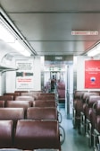 An empty train car with rows of brown leather seats lined uniformly. Large signs with public service announcements are posted on the walls, highlighting issues like opioid alternatives and real estate services. Overhead, fluorescent lighting provides illumination, and an emergency exit sign is visible on the ceiling.