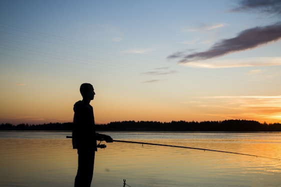 A serene lakeside scene with an angler holding a sturdy fishing rod from brgnyika llc, casting toward the sparkling water at dawn.