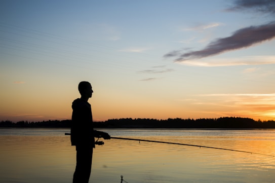 A silhouetted figure stands by a tranquil lake, holding a fishing rod. The background features a serene sunset with soft hues reflecting off the water's surface, creating a peaceful atmosphere.