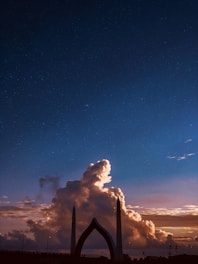 A dramatic scene featuring a towering bridge silhouette against a vast sky filled with large, illuminated cumulus clouds. The sky transitions from a deep blue starry night to a warm, golden sunset near the horizon.