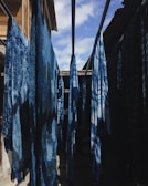 Several indigo-dyed fabrics hang on rods in an outdoor setting, with sunlight creating shadows on the cloths. The scene is framed by wooden and metal structures, with a partly cloudy sky visible above.