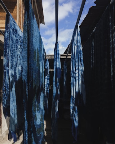 Several indigo-dyed fabrics hang on rods in an outdoor setting, with sunlight creating shadows on the cloths. The scene is framed by wooden and metal structures, with a partly cloudy sky visible above.