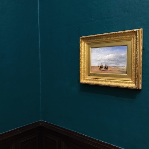 A small framed painting with a golden ornate frame is hung on a dark teal wall. The painting depicts a landscape with two figures on horseback in a vast, open area under a sky filled with white clouds. The wall and lighting suggest it is located in an art gallery or museum.