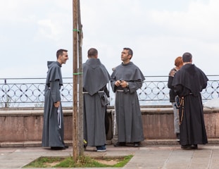 Several individuals dressed in long religious robes stand near a stone balustrade, engaged in conversation. One person appears to be laughing, while another holds a rosary. The background shows a cloudy sky.