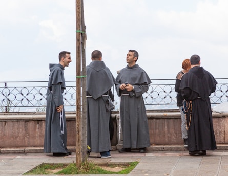 Several individuals dressed in long religious robes stand near a stone balustrade, engaged in conversation. One person appears to be laughing, while another holds a rosary. The background shows a cloudy sky.