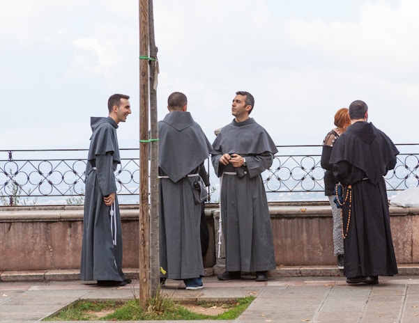 Several individuals dressed in long religious robes stand near a stone balustrade, engaged in conversation. One person appears to be laughing, while another holds a rosary. The background shows a cloudy sky.