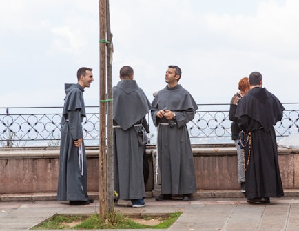 Several individuals dressed in long religious robes stand near a stone balustrade, engaged in conversation. One person appears to be laughing, while another holds a rosary. The background shows a cloudy sky.