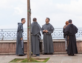 Several individuals dressed in long religious robes stand near a stone balustrade, engaged in conversation. One person appears to be laughing, while another holds a rosary. The background shows a cloudy sky.