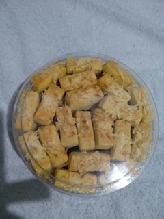 A plastic container filled with rectangular pastries topped with shredded cheese, resting on a light-colored fabric background.