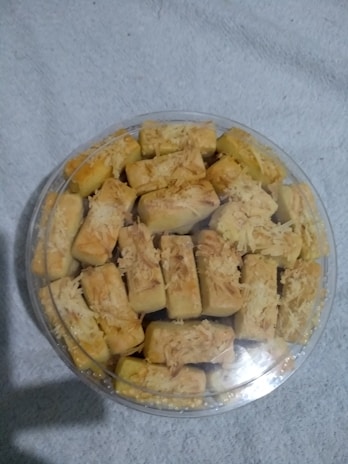 A plastic container filled with rectangular pastries topped with shredded cheese, resting on a light-colored fabric background.