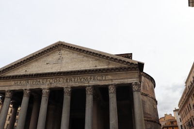 An ancient Roman building with large Corinthian columns supporting a pediment. The inscription 'M.AGRIPPA.L.F.COSTERTIUM.FECIT' is visible on the facade. The surrounding structures are part of a historical urban setting.