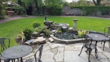 A tranquil garden features a well-maintained lawn surrounded by various plants and shrubs. In the foreground, metal patio tables and chairs are placed on a stone-paved area. A small pond with a waterfall and decorative stone border is centrally located. A birdbath stands next to the pond, and a garden wall covered with climbing plants is visible in the background.