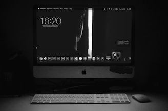 A dark room is illuminated by the glow of a computer screen displaying a minimalist desktop interface. The screen shows the time and date, along with various application icons. A wireless keyboard and mouse are positioned in front of the monitor. The scene is primarily in black and white, emphasizing the contrast between light and shadow.