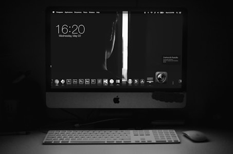 A dark room is illuminated by the glow of a computer screen displaying a minimalist desktop interface. The screen shows the time and date, along with various application icons. A wireless keyboard and mouse are positioned in front of the monitor. The scene is primarily in black and white, emphasizing the contrast between light and shadow.