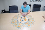A man in a light blue shirt is seated at a large conference table, which has a colorful circular design featuring an intricate pattern and the word 'Kaleidico' in the center. The table is set in a modern office environment with black chairs, a wall with mirrors, and electronic equipment in the background.